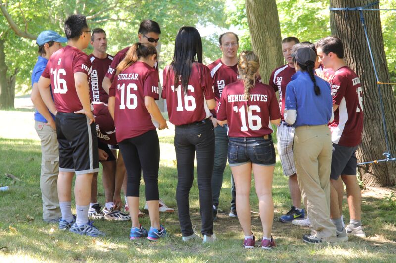 The image shows a group of people in maroon shirts with the number 16 on them, standing in a circle outdoors. They appear to be a team or group of friends. Some are wearing shorts, while others have pants or leggings. The setting is a park or outdoor area with trees.